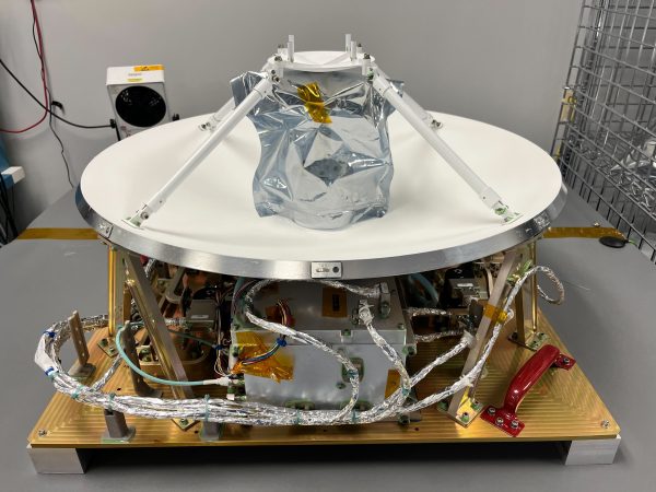 The Polylingual Experimental Terminal is the focus of this photograph. We see a white antenna dish, approximately 0.6-meters in size, facing the ceiling, sitting on a golden platform. Silver wires resembling foil are shown protruding beneath the antenna dish. The terminal sits on top of a grey table inside a white laboratory.