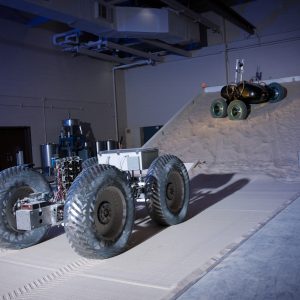 Two test vehicles driving on GRC-1 lunar simulant in the SLOPE Lab; one on a flat surface and one on the adjustable tilt-bed