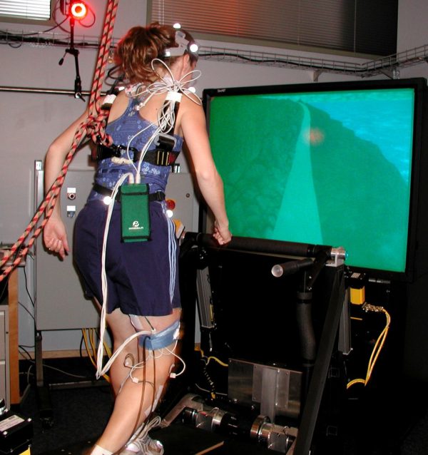 A women with wires and equipment attached to her body walks on treadmill in front of a TV.